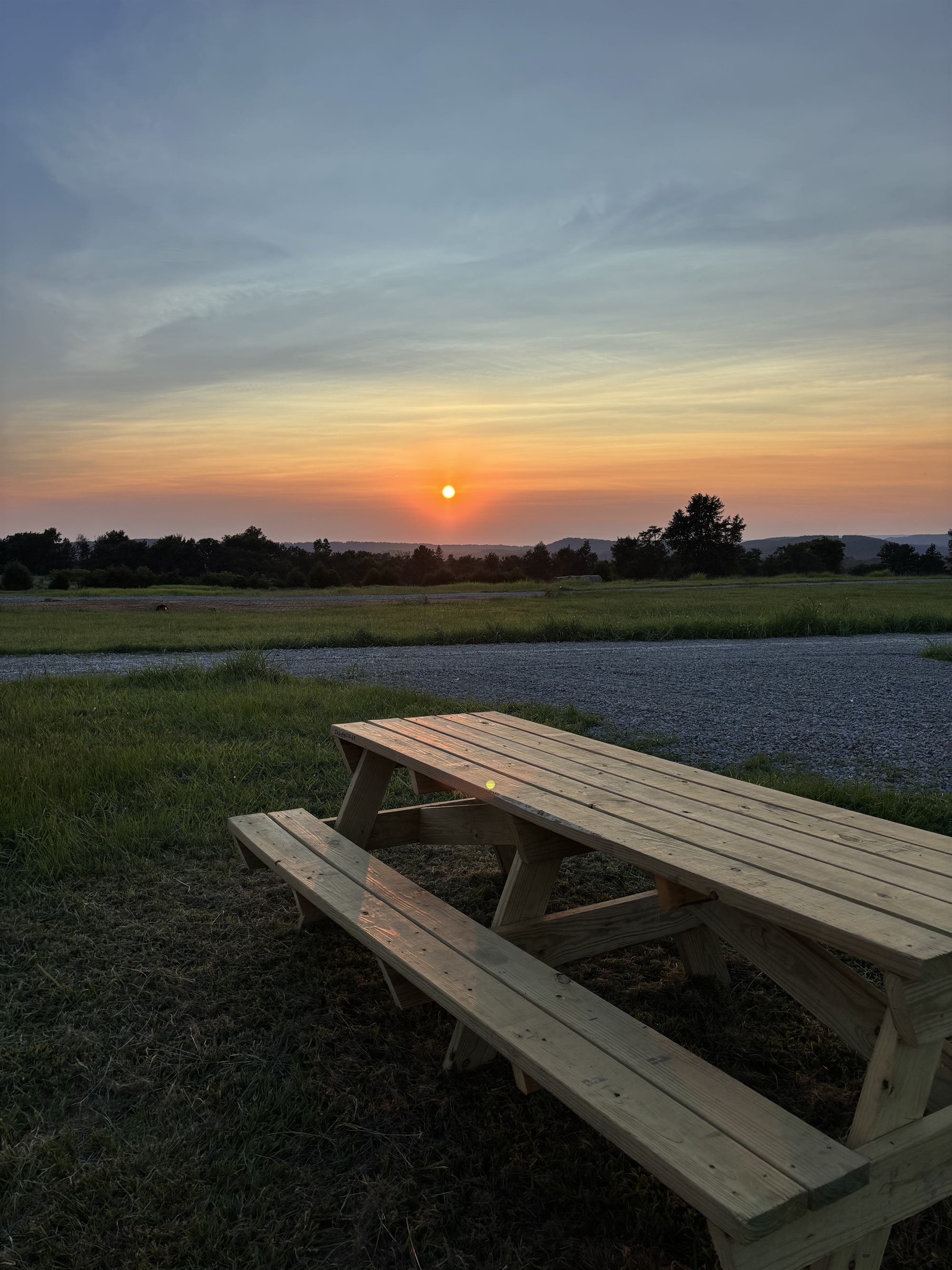 Sunset over the rolling hills at The Pineys RV Park, with a new wooden picnic table beside the gravel loop drive